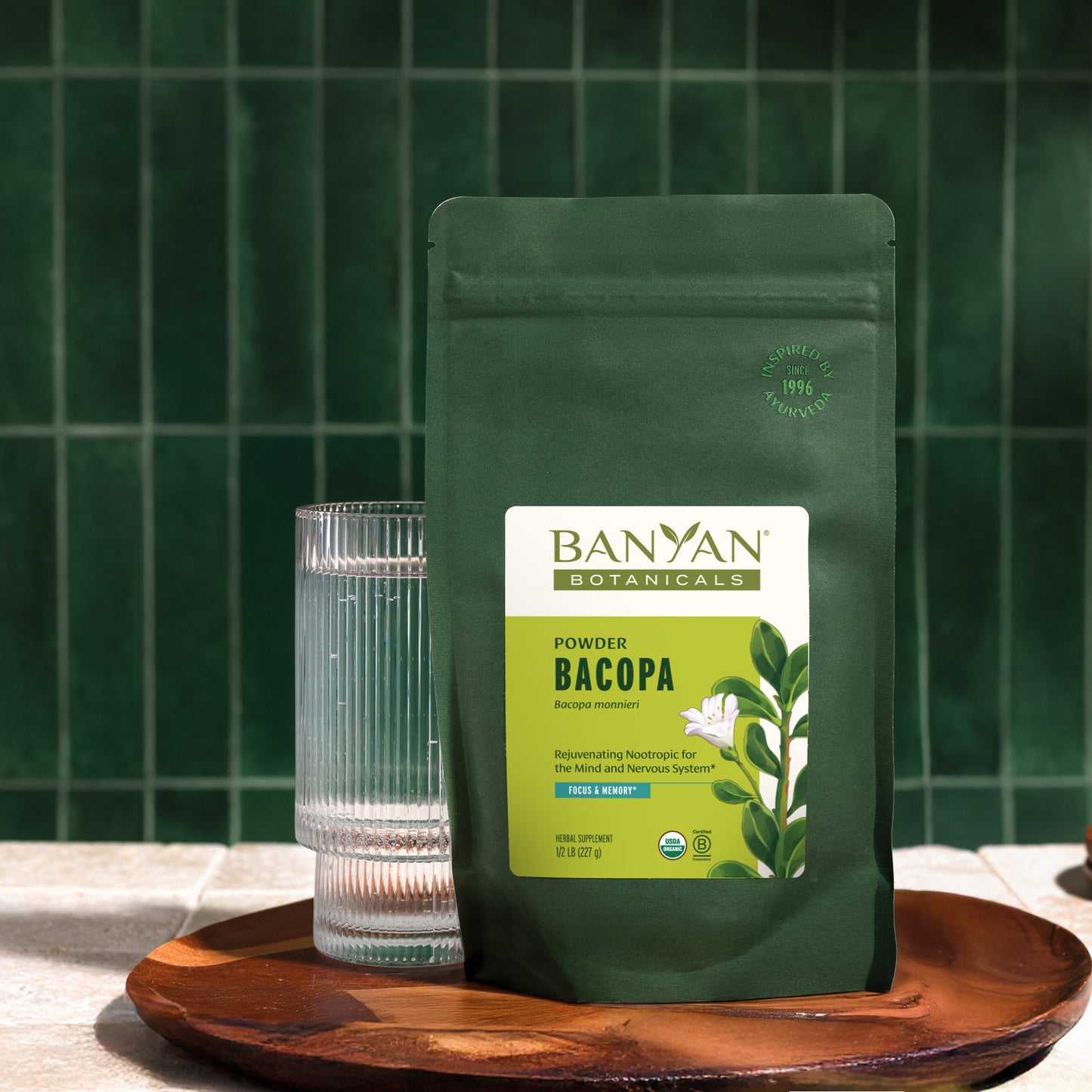 1/2 lb: Banyan Botanicals Bacopa Powder pouch with a glass of water on a wooden tray against a green tiled wall.