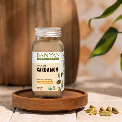 spice jar: Glass spice jar of Banyan Botanicals Organic Cardamom on a wooden tray with cardamom pods.