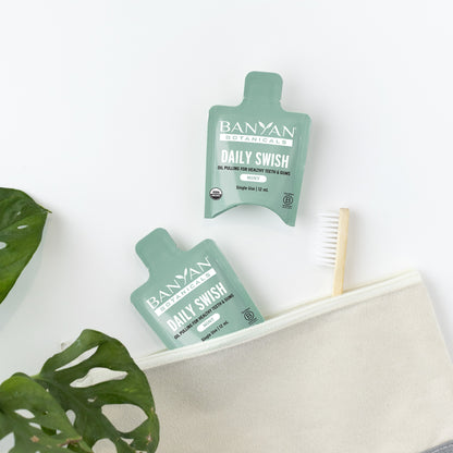 Single Use: Two green packets of Banyan Botanicals single use Daily Swish on a white surface with a plant leaf and toothbrush in the background.